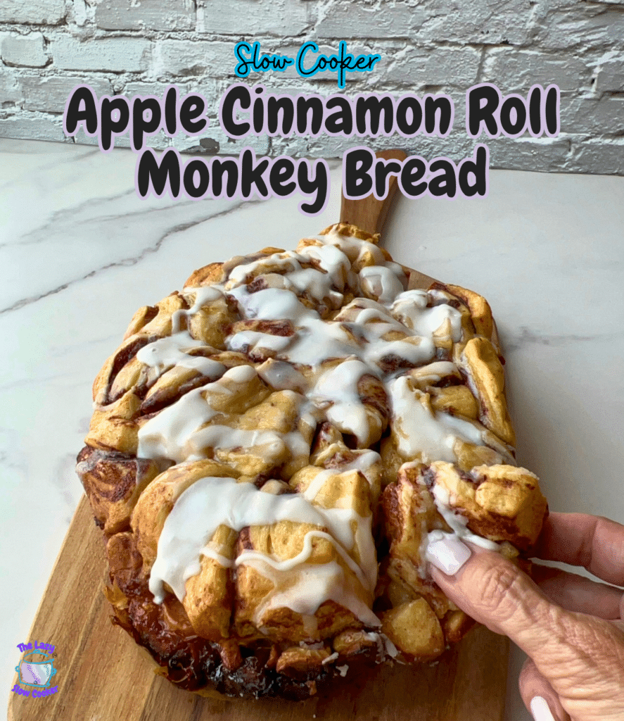 A hand lifts a piece of apple cinnamon roll monkey bread from a wooden board. The words “Slow Cooker Apple Cinnamon Roll Monkey Bread” appear at the top of the image.