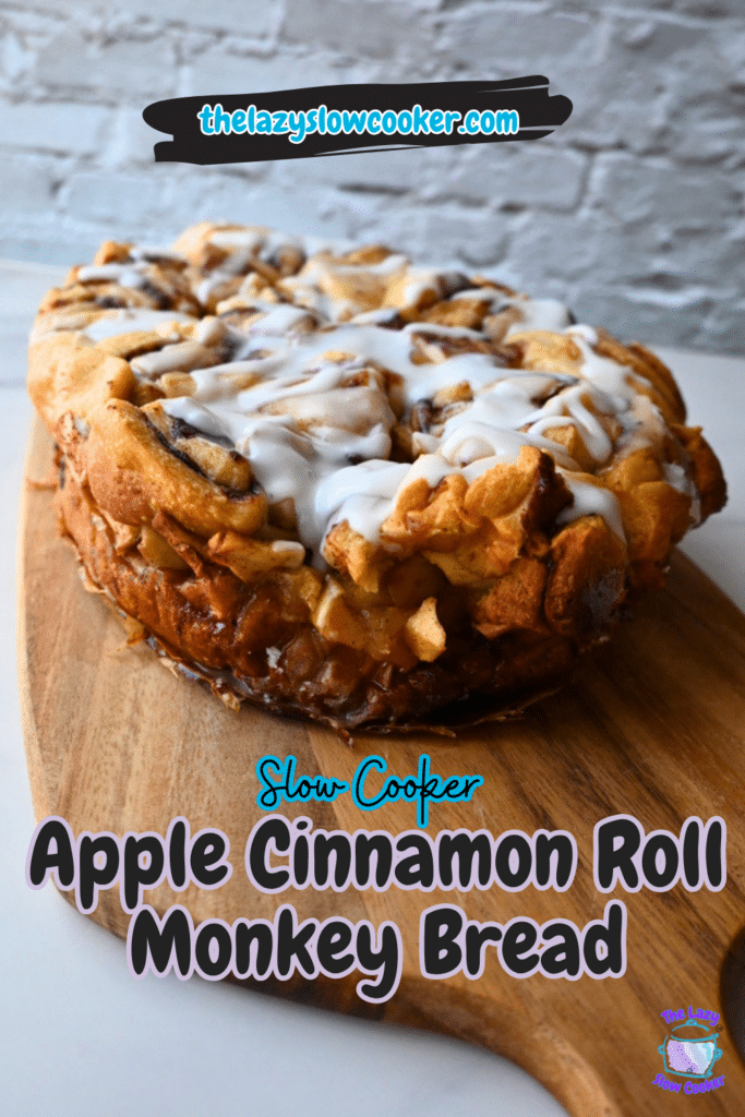 A loaf of apple cinnamon roll monkey bread drizzled with white icing rests on a wooden board. The image includes the recipe name, “Slow Cooker Apple Monkey Bread,” and the website “thelazyslowcooker.com” displayed over a light brick background.