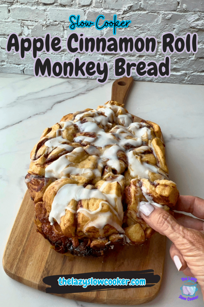A hand breaks off a piece of apple cinnamon roll monkey bread on a wooden board. The recipe name, “Slow Cooker Apple Cinnamon Roll Monkey Bread,” and the website “thelazy slowcooker.com” are shown in the image.