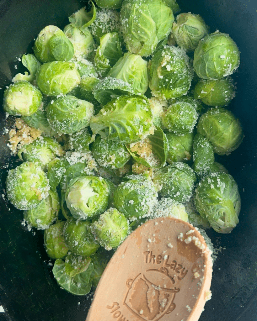 Cooked Brussels sprouts in a slow cooker, topped with cheese and garlic, with a wooden spoon labeled The Lazy Slow Cooker beside them.