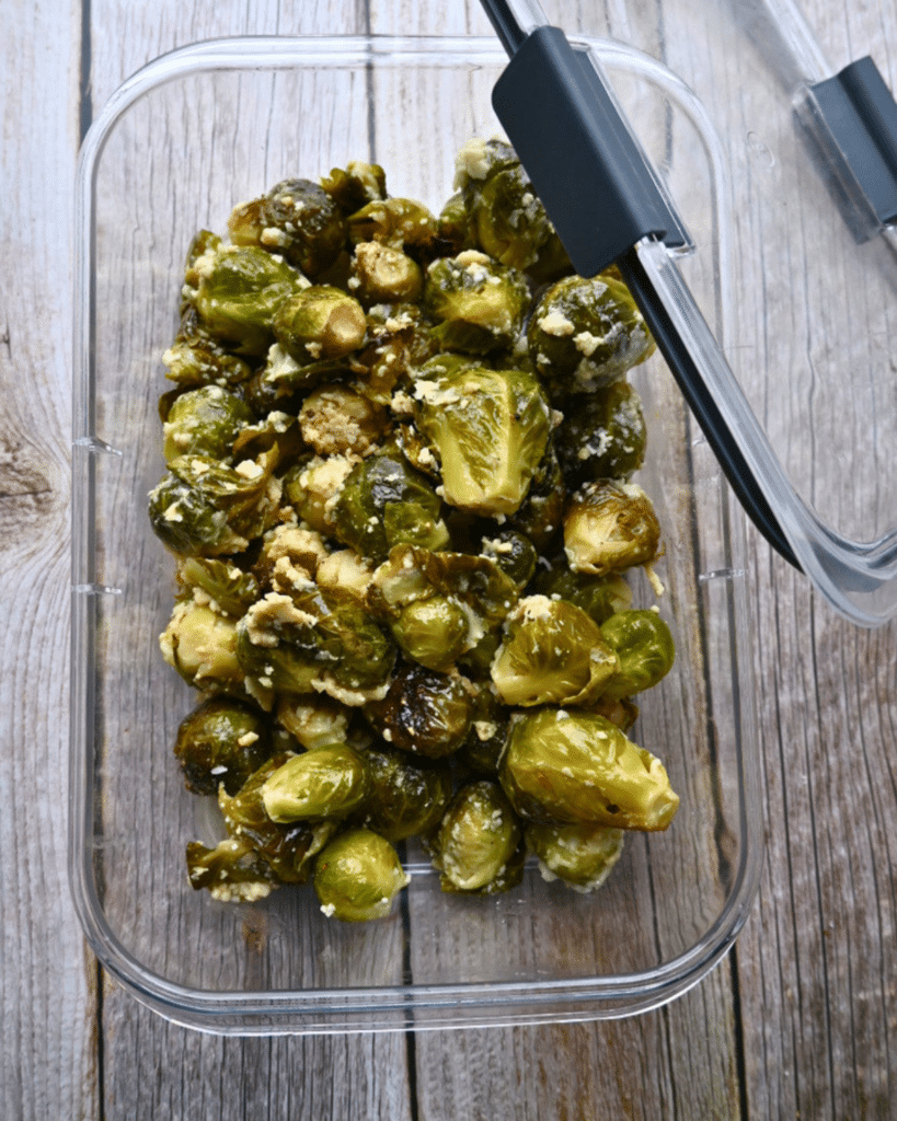 A clear container with slow cooker Brussels sprouts topped with crumbled cheese rests on wood, its lid slightly open.