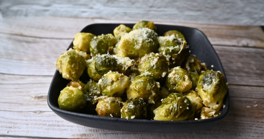 A black square bowl holds roasted Brussels sprouts cooked in a slow cooker, topped with grated Parmesan, on a light wood table.