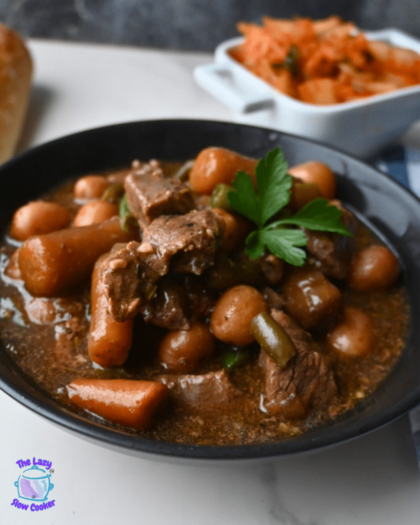 A black bowl holds garlic herb beef stew with potatoes and carrots, topped with parsley. Kimchi, bread, and The Lazy Slow Cooker logo appear behind.