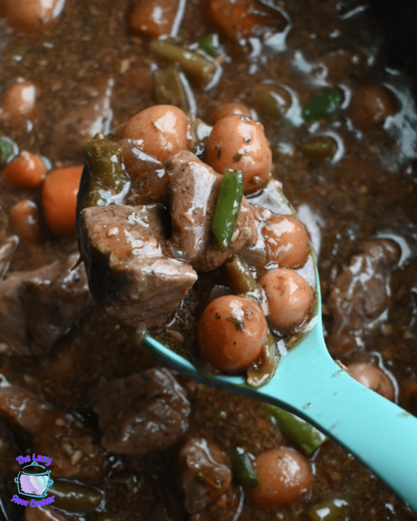 A spoon holds a serving of slow cooker garlic herb beef stew with beef, carrots, green beans, and potatoes above the pot.