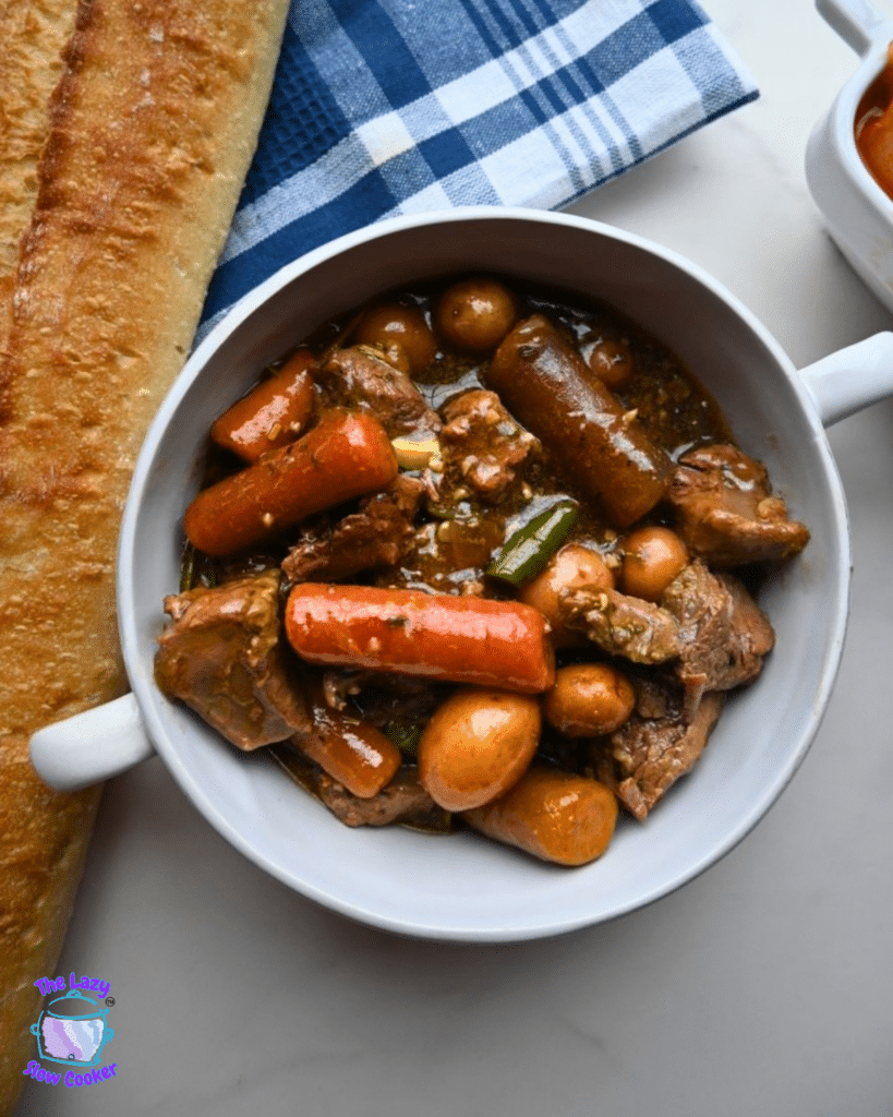 A white bowl of slow cooker garlic herb beef stew sits beside a baguette and blue-and-white checkered napkin on a white table.