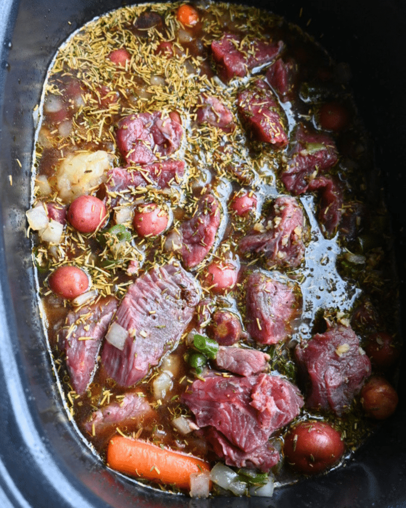 Raw beef chunks, whole baby potatoes, carrots, onions, and herbs sit in broth inside a slow cooker before becoming beef stew.
