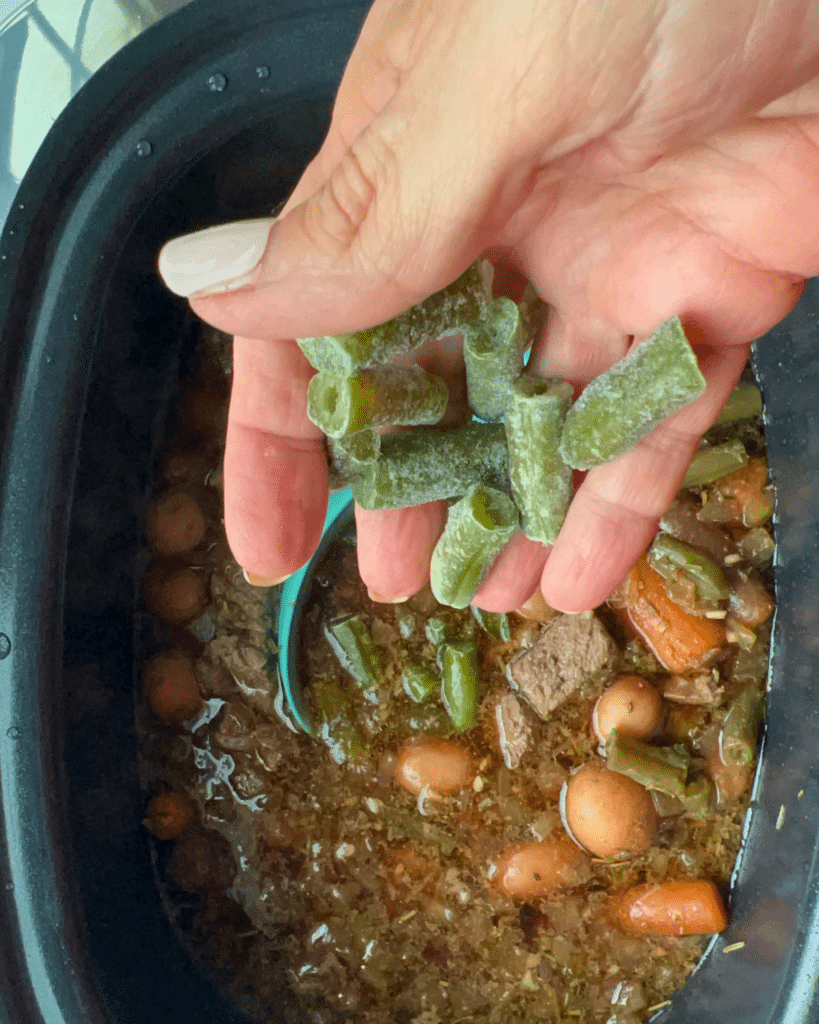 A hand adds frozen green beans to a slow cooker filled with garlic herb beef stew, including chunks of beef, potatoes, and carrots.
