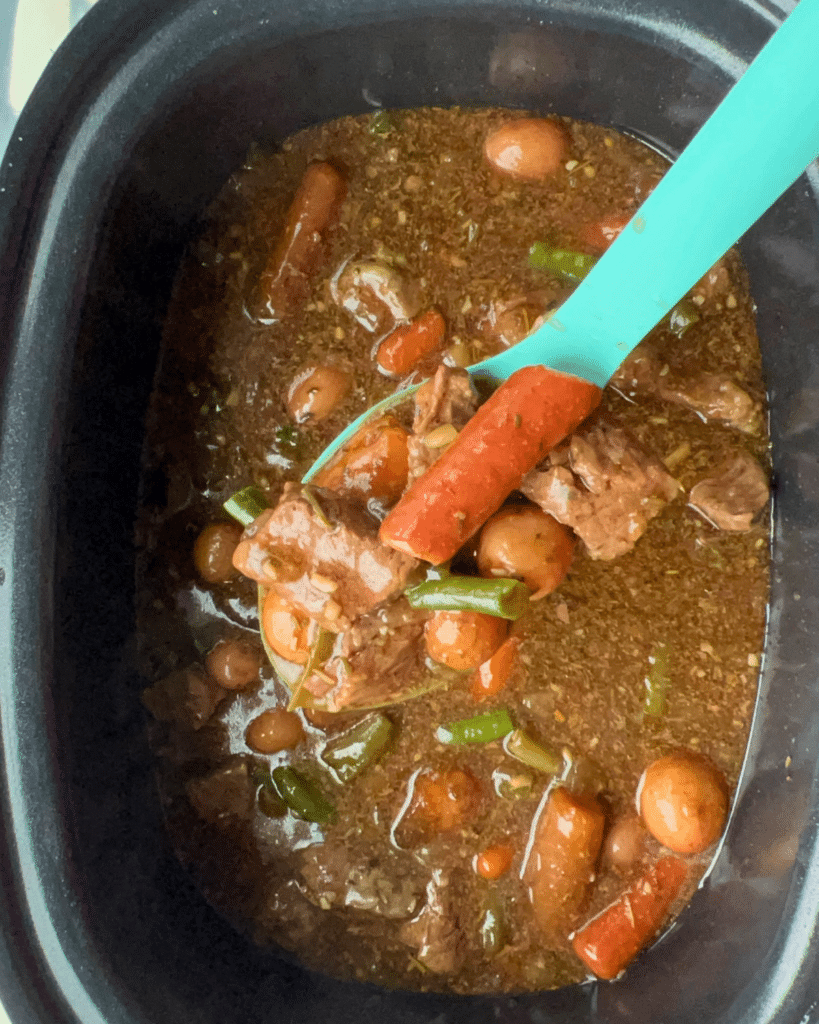 A slow cooker of garlic herb beef stew with visible beef, carrots, potatoes, and green beans; a blue spoon lifts some stew.