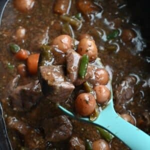 A blue ladle lifts garlic herb beef stew from a pot, showing beef, potatoes, carrots, green beans, and herbs in rich brown broth.