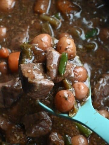 A blue ladle lifts garlic herb beef stew from a pot, showing beef, potatoes, carrots, green beans, and herbs in rich brown broth.