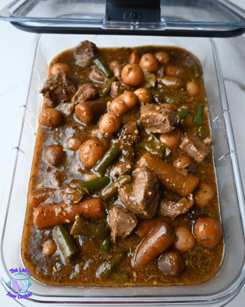 A clear container of slow cooker garlic herb beef stew with visible beef, potatoes, carrots, green beans, and onions in brown broth.