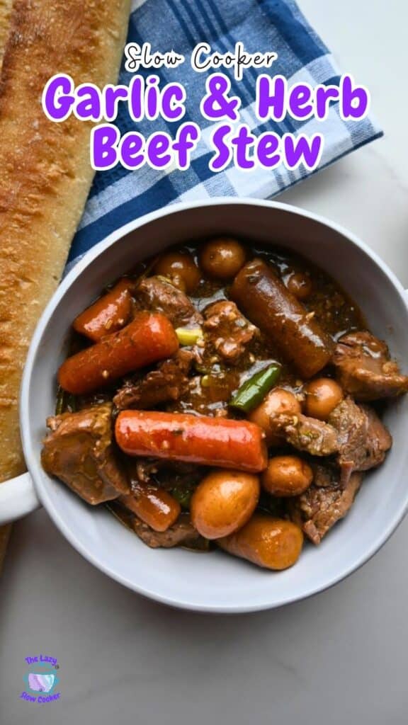 A bowl of garlic herb beef stew with carrots and potatoes is on a white surface beside a baguette and blue-striped napkin. Recipe title above.