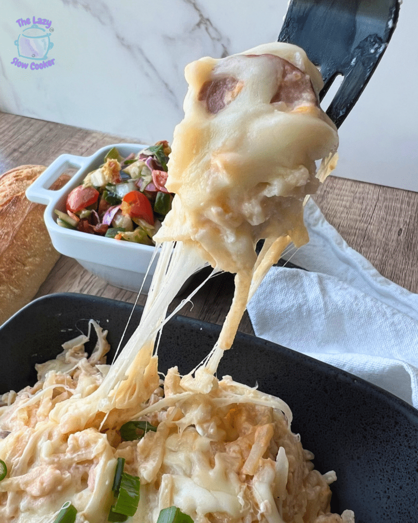 A spatula lifts a cheesy serving of slow cooker kielbasa and potato casserole from a black dish, with cheese stretching. Bread, salad, and a napkin are on the wooden table behind.