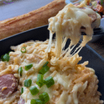 Close-up of slow cooker kielbasa and potato casserole with sausage, cheese, and green onions. A fork lifts a cheesy bite; sides behind.