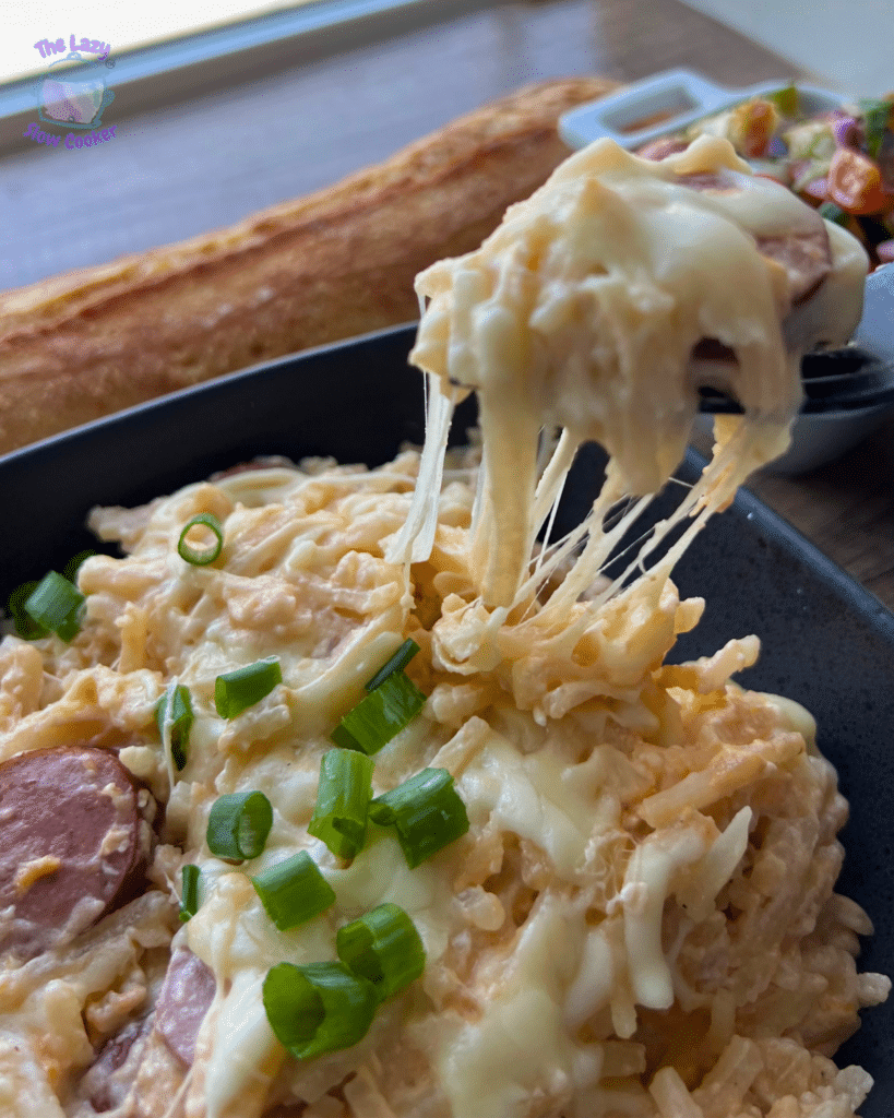 Close-up of slow cooker kielbasa and potato casserole with sausage, cheese, and green onions. A fork lifts a cheesy bite; sides behind.