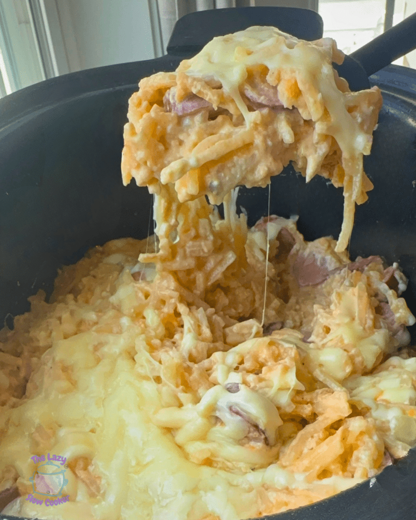 A spoon lifts a portion of creamy, cheesy casserole from a pan, showing stretchy cheese, ham pieces, and shredded potatoes.