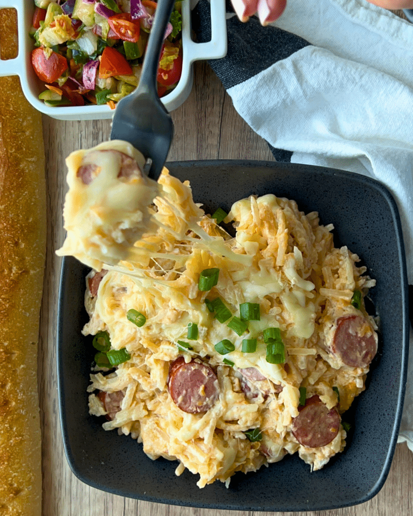 A black bowl holds cheesy slow cooker kielbasa and potato casserole with sausage and green onions. A fork lifts a serving. Salad and bread are beside it on a wooden table with a white napkin.