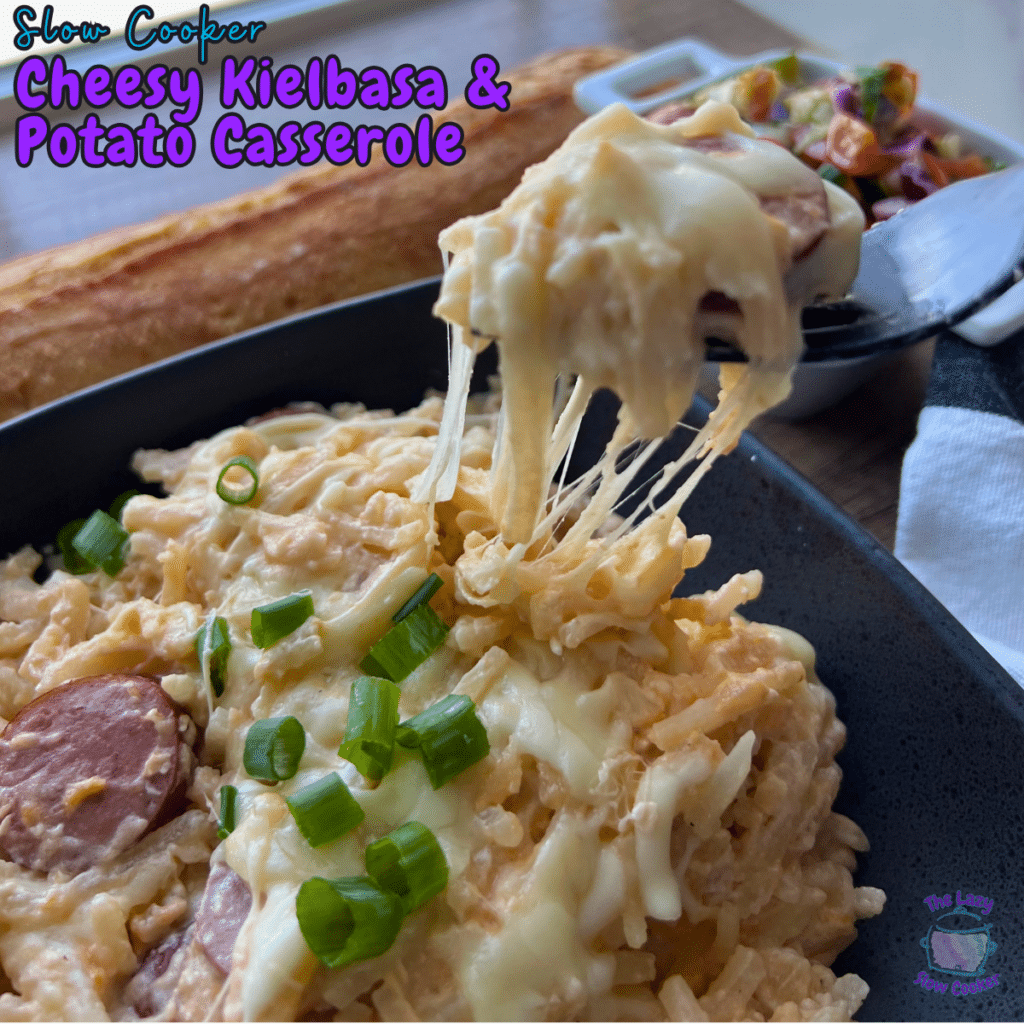 A fork lifts a cheesy serving of slow cooker kielbasa and potato casserole from a black dish, with salad and breadstick behind.