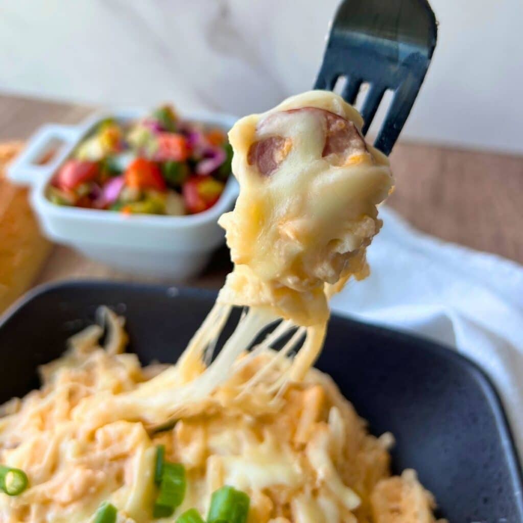A fork lifts cheesy pasta with sausage from a bowl of slow cooker kielbasa and potatoes; salad sits in a white dish behind.