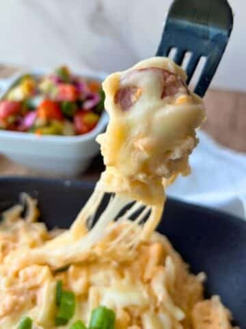 A fork lifts cheesy pasta with sausage from a bowl of slow cooker kielbasa and potatoes; salad sits in a white dish behind.