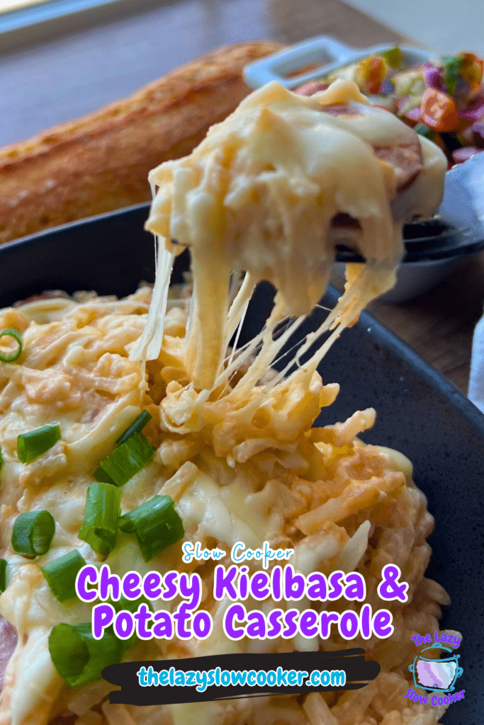 A spoon lifts a cheesy portion of slow cooker kielbasa and potato casserole, topped with green onions. Breadstick and salad behind.