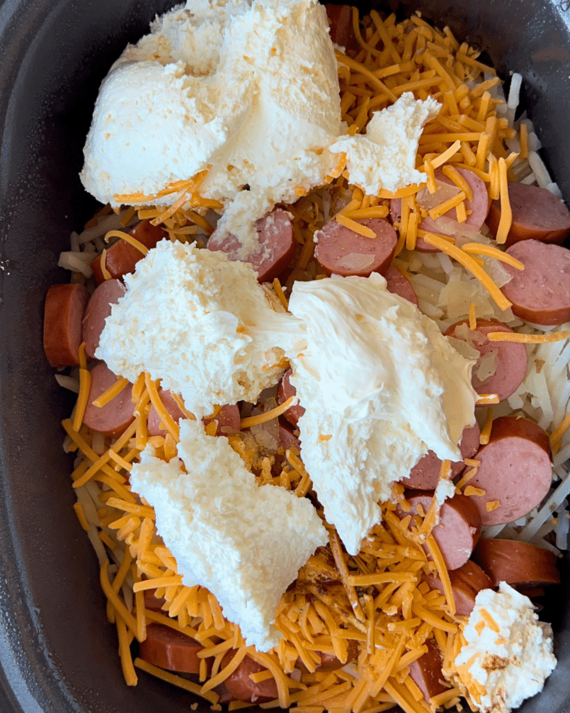 In a slow cooker, sliced kielbasa, shredded cheddar, and cream cheese are layered on top of hash browns before cooking.