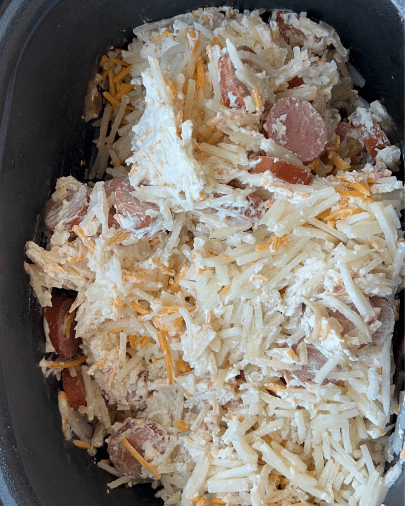 A slow cooker filled with layers of shredded cheese, hash browns, sliced kielbasa, and creamy sauce before cooking.