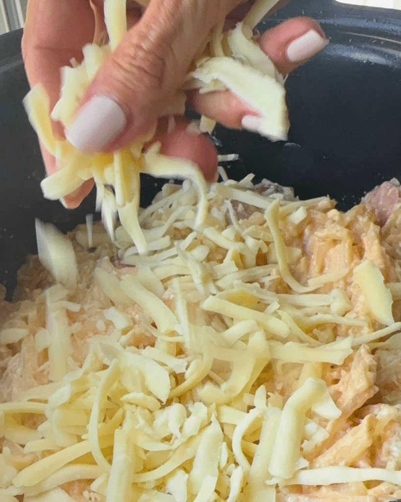 A hand with pale pink nails sprinkles cheese over creamy kielbasa and potatoes in a black slow cooker.