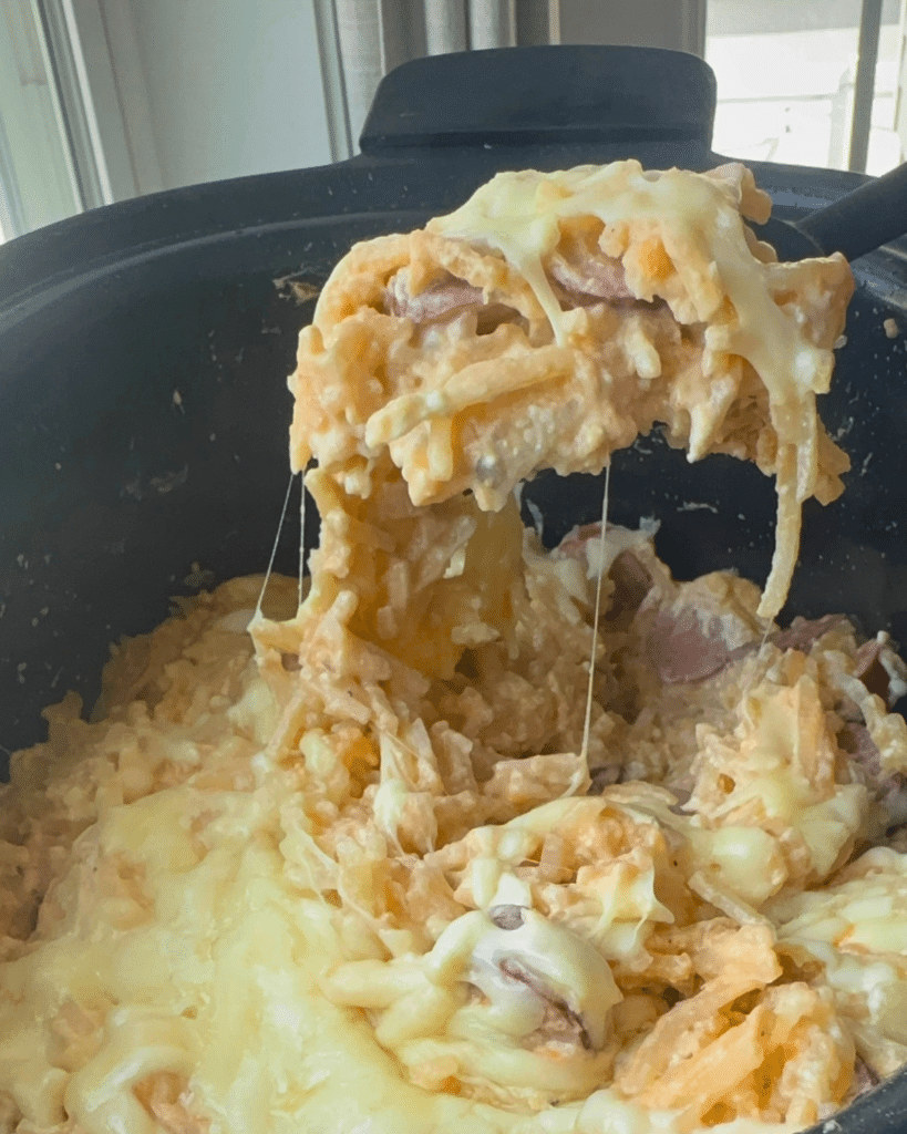 A scoop lifts cheesy kielbasa and potato casserole from a black slow cooker, with gooey cheese stretching over tender potatoes.