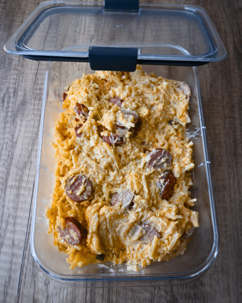 A glass dish of creamy sausage, potato, and shredded cheese mixture rests on a wooden table with its lid slightly ajar.