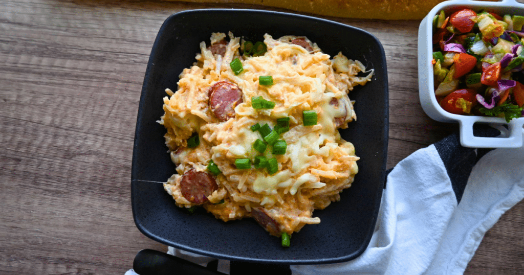 Cheesy hash brown casserole with kielbasa and green onions in a black bowl, beside a vibrant side salad on a wooden table.
