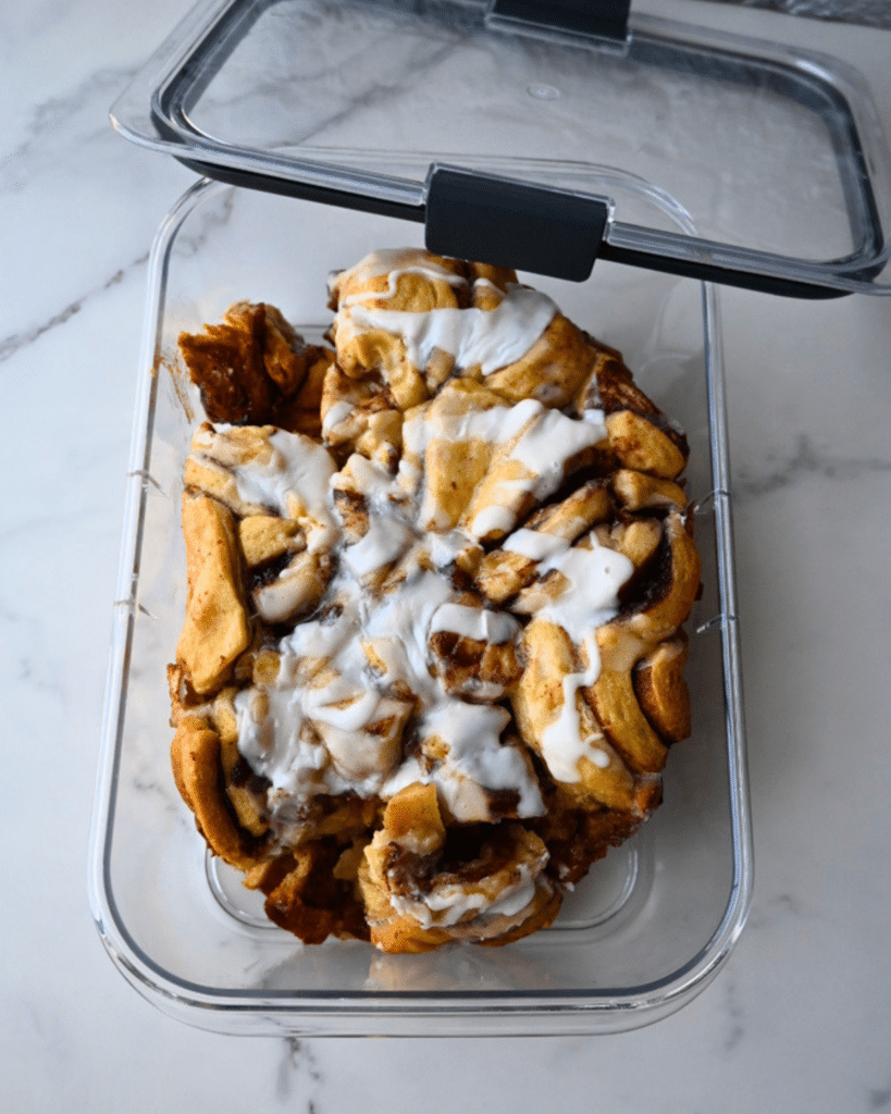 A clear, rectangular plastic container with the lid partly open holds iced cinnamon rolls and broken pieces of slow cooker apple monkey bread. The baked goods look slightly messy, and the container sits on a white marble counter.