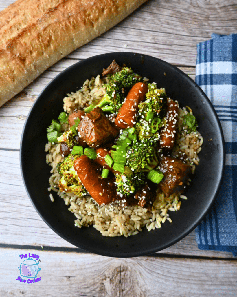 A black bowl of slow cooker soy ginger beef with rice, carrots, and broccoli, garnished with sesame seeds and green onions. Bread and a blue plaid napkin are beside the bowl on a light wood table.