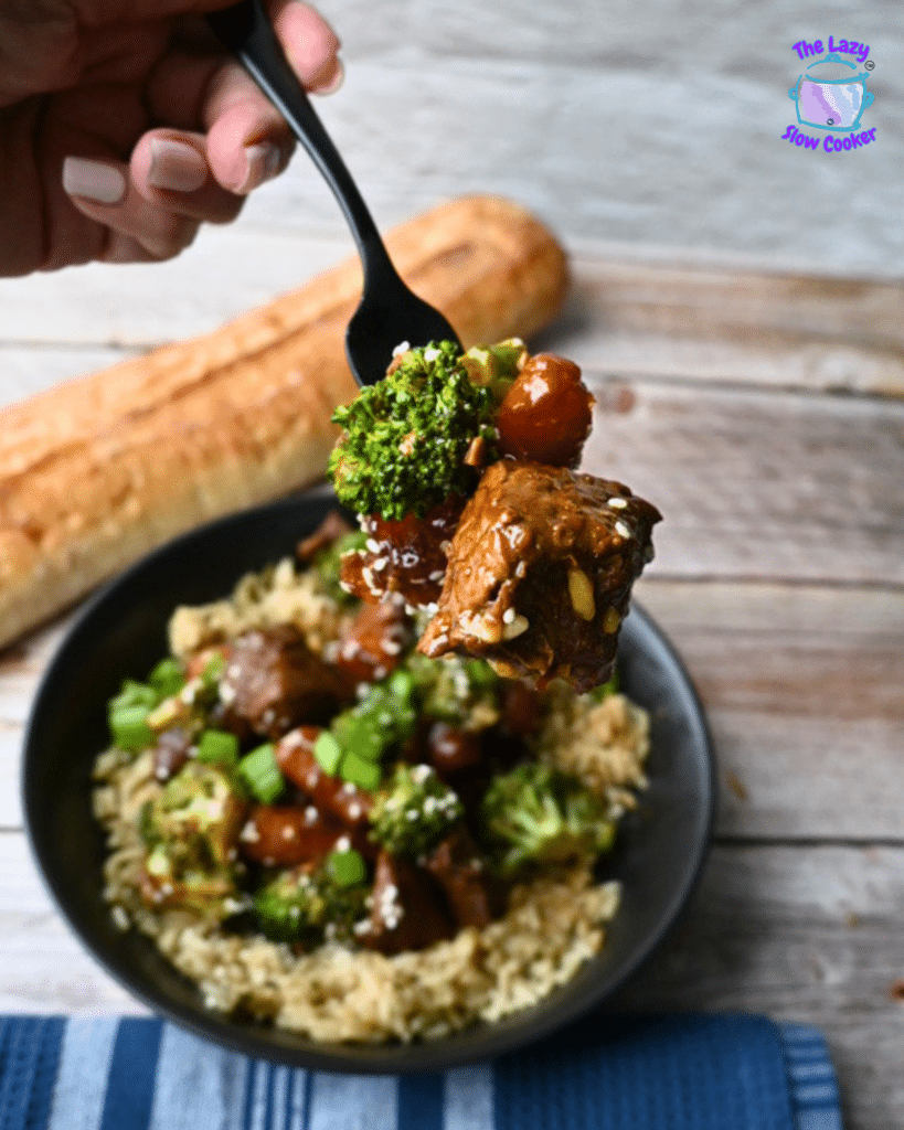 A hand holds a fork with soy ginger beef, broccoli, and veggies over a rice bowl. A baguette and The Lazy Slow Cooker logo are visible.