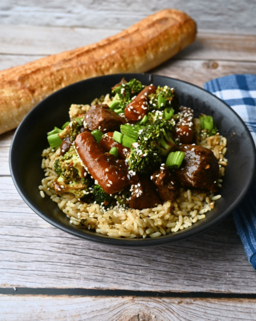 A black bowl holds rice with slow cooker soy ginger beef, broccoli, carrots, green onions, and sesame seeds on a wooden table.