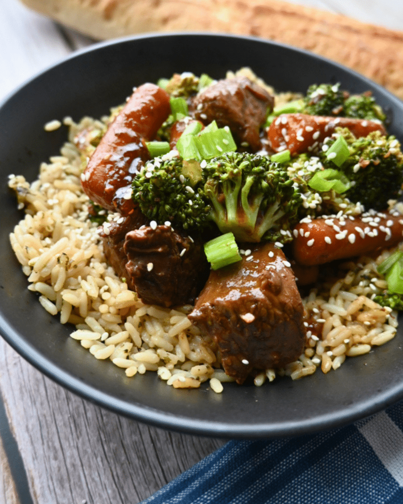 A black bowl holds rice, slow cooker soy ginger beef, broccoli, and carrots, topped with sesame seeds and green onions. Bread sits behind.