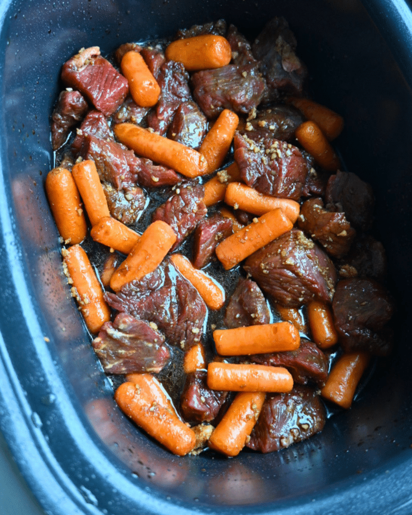 Raw beef chunks and baby carrots sit in a black slow cooker, coated in a soy ginger marinade, ready for cooking.