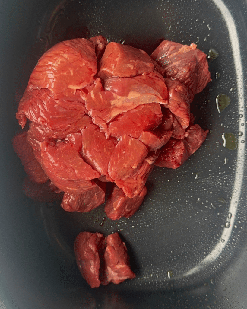 Several raw pieces of red beef sit in a black pan with a thin layer of oil, ready to start slow cooker soy ginger beef.