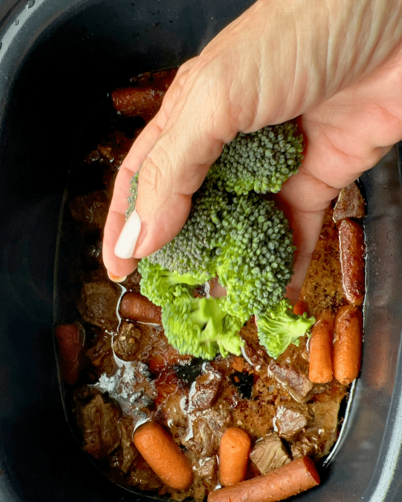 A hand holds broccoli florets over a slow cooker with soy ginger beef, carrots, and broth, about to add the broccoli to the dish.