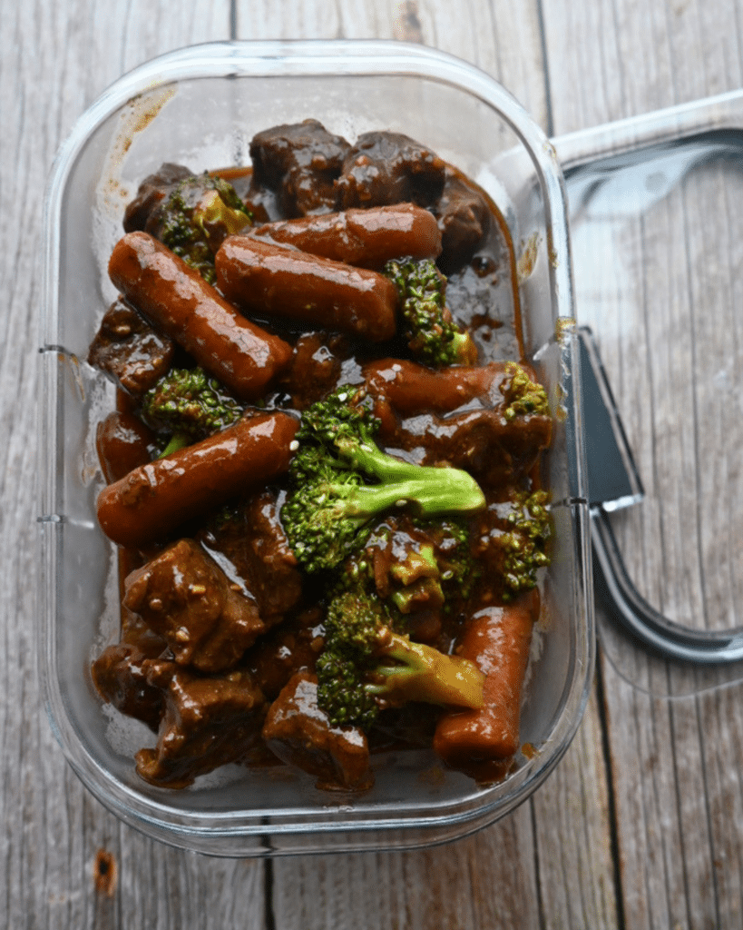 A glass container of slow cooker soy ginger beef stew with carrots and broccoli sits on wood, the lid slightly ajar.