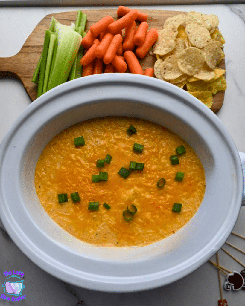 A white slow cooker holds buffalo chicken dip with melted cheese and ground chicken, topped with chopped green onions. Celery sticks, baby carrots, and tortilla chips are arranged around the cooker on a wooden board.