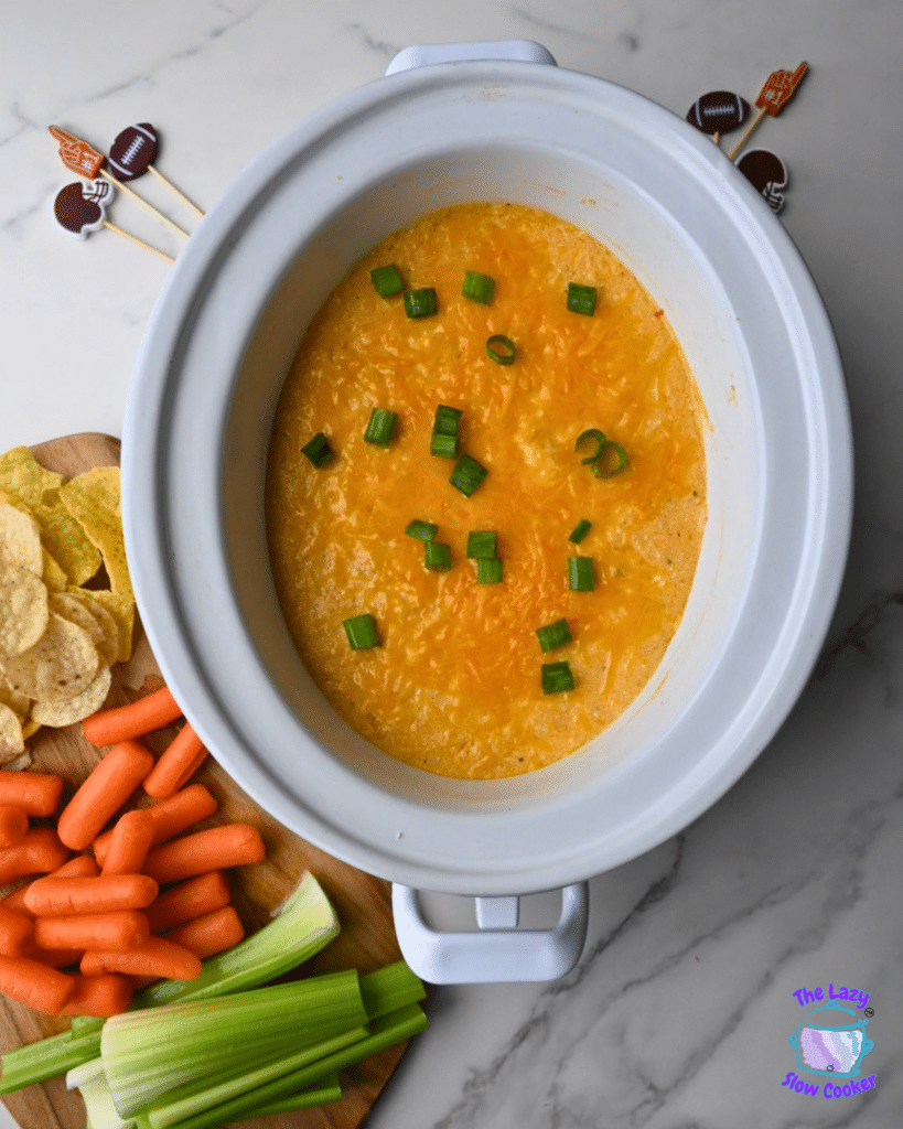 A white slow cooker holds ground chicken buffalo dip covered in melted cheese and chopped green onions. Next to the slow cooker are tortilla chips, baby carrots, celery sticks, and a few football-themed party picks on a marble countertop.