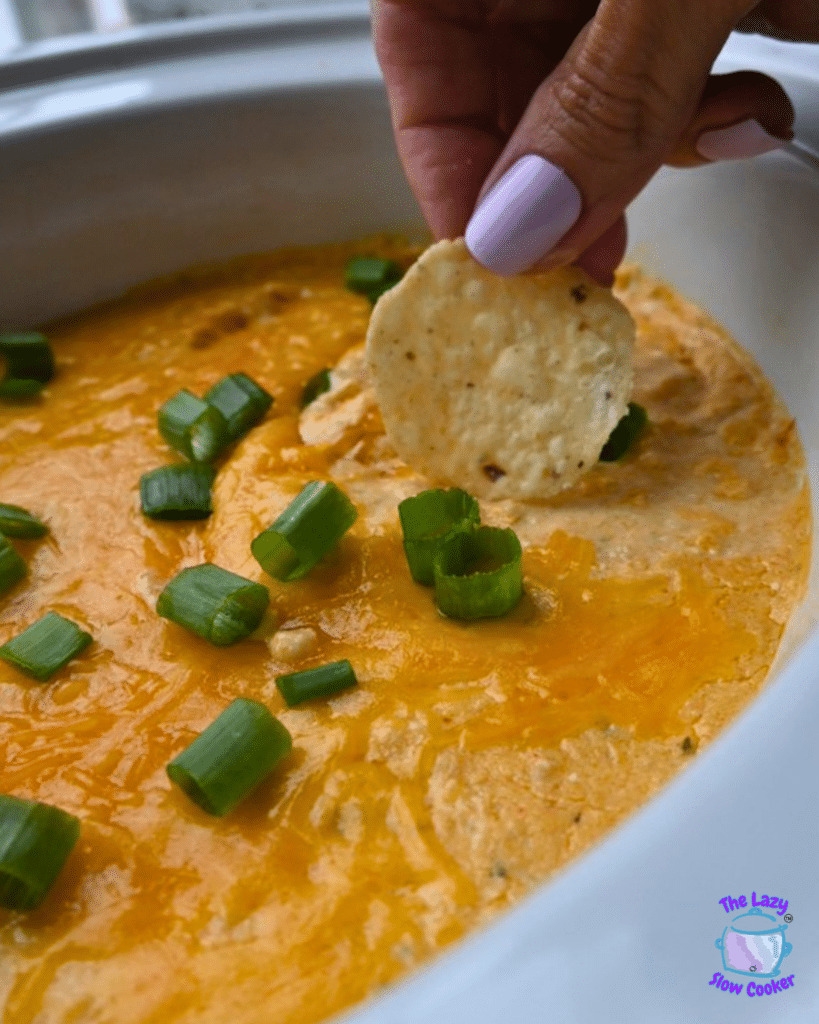 A hand with painted nails dips a tortilla chip into a creamy buffalo ground chicken dip, topped with green onions and served in a white dish. The Lazy Slow Cooker logo appears in the corner.