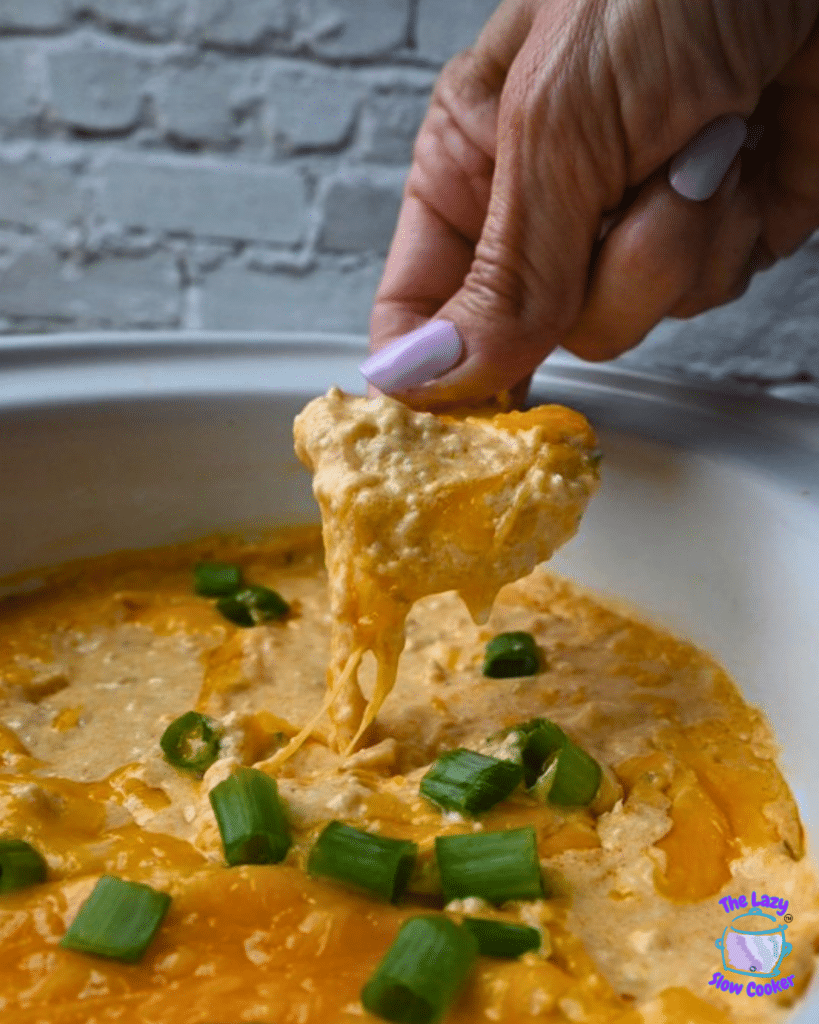 A person with lavender-painted nails dips a chip into a white dish of cheesy ground chicken buffalo dip, garnished with green onions. The dish sits in front of a white brick background.