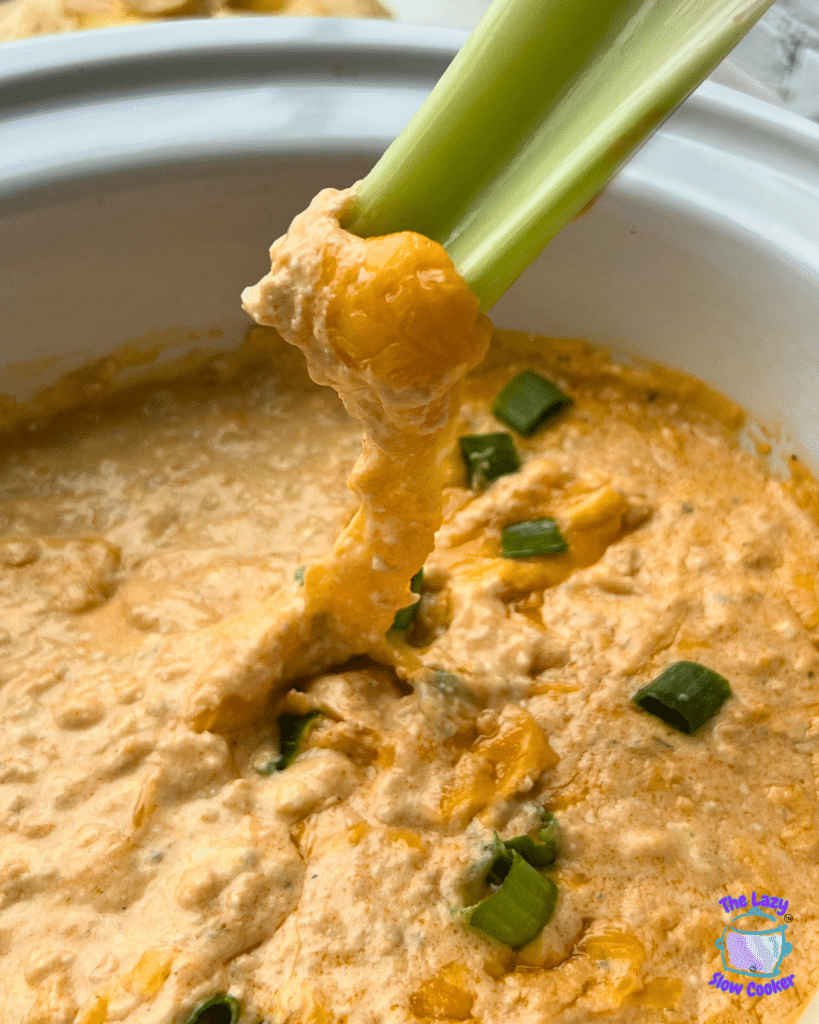 A hand dips a piece of celery into a bowl of creamy orange buffalo chicken dip topped with melted cheese and sliced green onions.