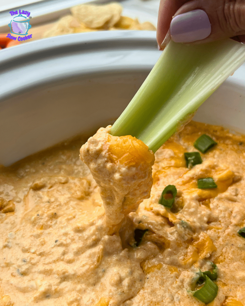 A hand dips a celery stick into creamy buffalo chicken dip, topped with chopped green onions, in a white dish. Blurry chips sit in the background.