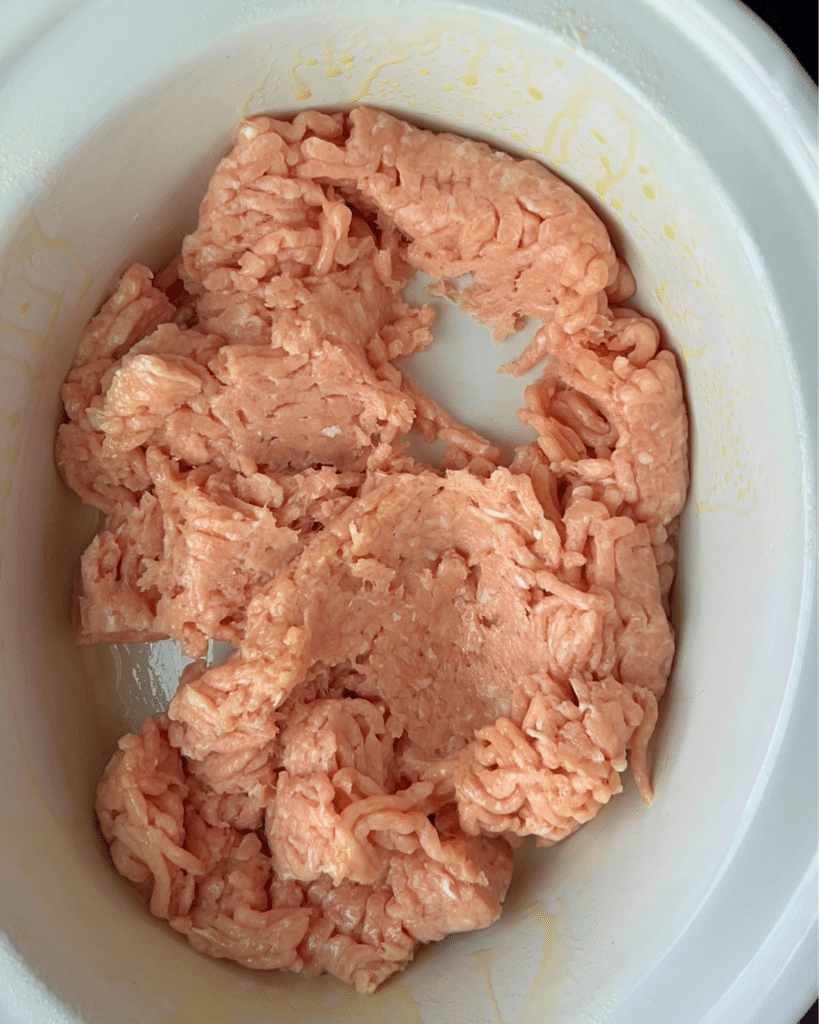 A white bowl filled with raw ground chicken sits in a slow cooker, ready to be used in a ground chicken buffalo chicken dip recipe.