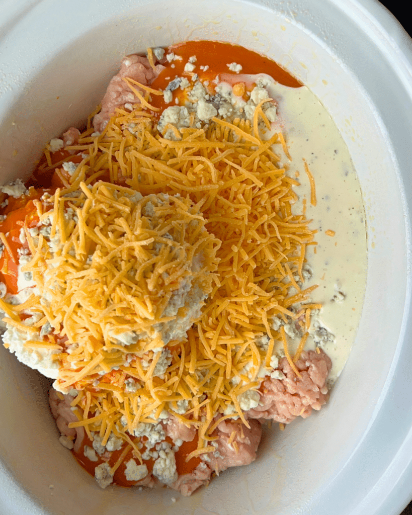 Inside a slow cooker, uncooked ground chicken is topped with piles of shredded cheddar cheese, blue cheese crumbles, buffalo sauce, and a swirl of creamy dressing, all about to be combined and cooked into buffalo chicken dip.