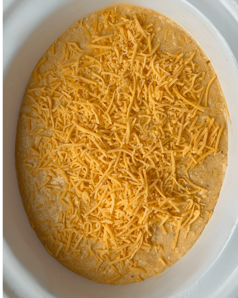 Shredded cheddar cheese covers a layer of ground chicken buffalo dip in a white oval baking dish, ready to be cooked.