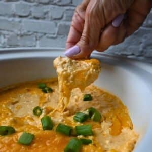 A person with lavender-painted nails dips a tortilla chip into a serving dish of cheesy ground chicken buffalo dip, which is topped with chopped green onions. The white dish sits in front of a white brick wall.
